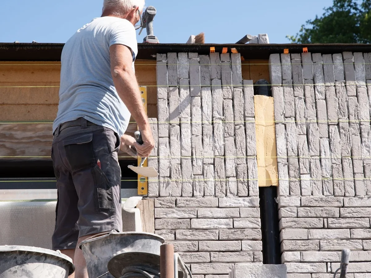 Mason laying courses of light colored brick against wall sheathing, brick veneer
