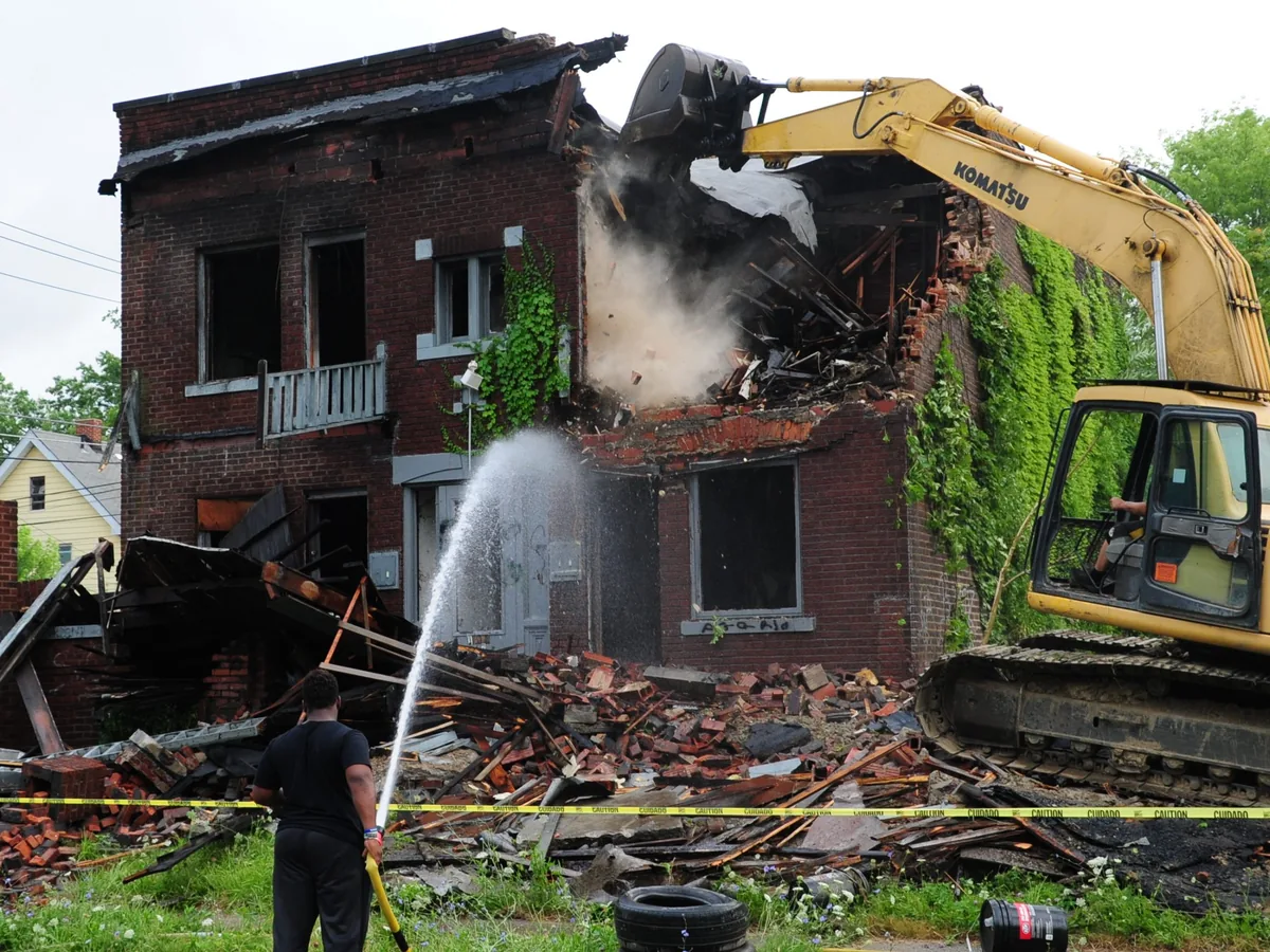 Excavator breaking down a two story brick building while a worker hoses down the dust, building demolition