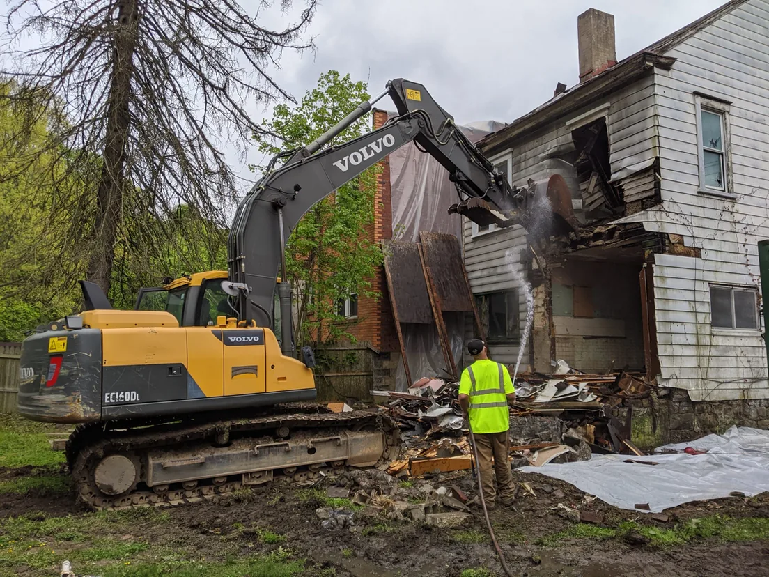 Excavator tearing into a two story wood frame house while a worker sprays water on the dust, house demolition