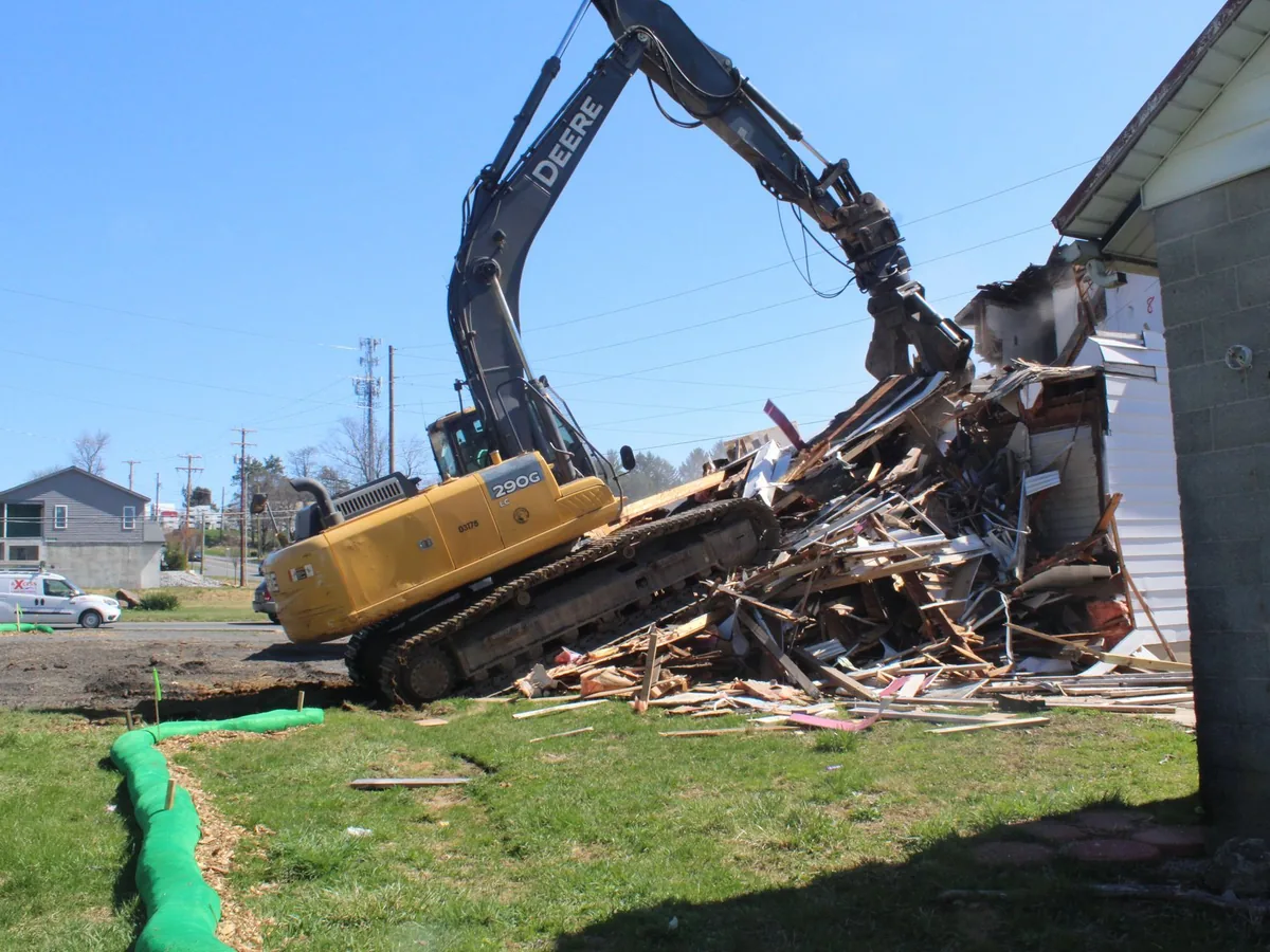 Excavator with a grapple pulling a house down into a debris pile beside a block garage, house demolition