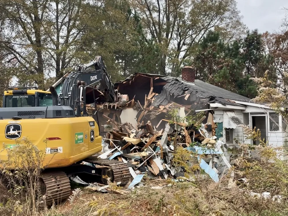 Excavator tearing down a small one story house among autumn trees, house demolition